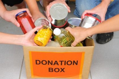 Hands placing canned and jarred food into a cardboard donation box labeled “DONATION BOX.”