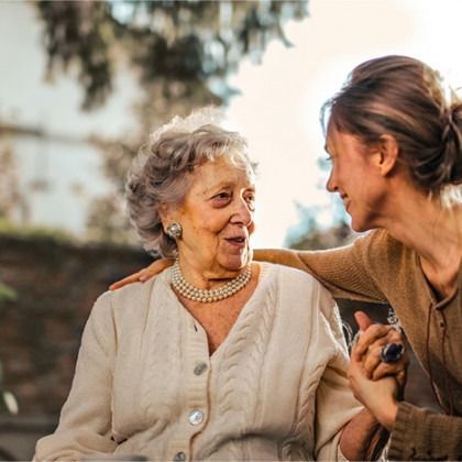 Two women smiling and talking outdoors, one gently touching the other's shoulder
