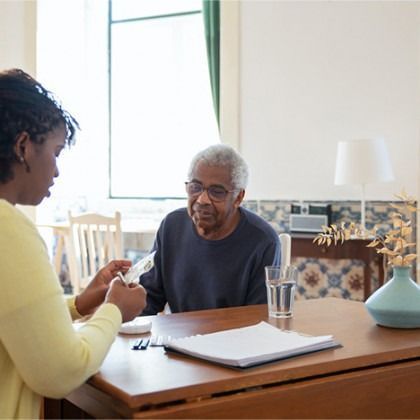 Two people talking at a table in a bright room, with papers, a glass of water, and a lamp.