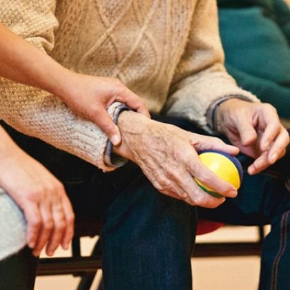 Older hands holding a yellow stress ball while seated beside another person on a couch