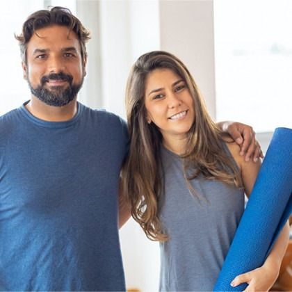 Two smiling people standing indoors, holding a blue yoga mat