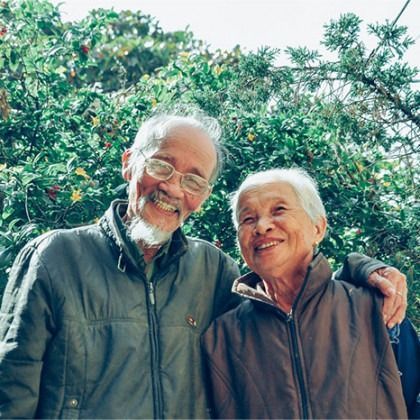 Two smiling people standing close together outdoors with lush green foliage behind them
