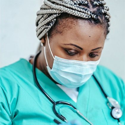 Healthcare worker in teal scrubs wearing a mask and stethoscope, looking down attentively.