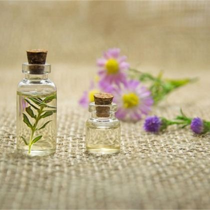 Two glass bottles with corks on burlap, filled with herbs and clear liquid, with purple flowers behind them
