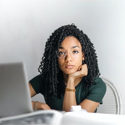 Woman with glasses resting her chin at a desk beside a laptop, looking at the camera.