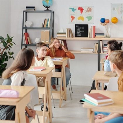 Students seated in a bright classroom, listening to a teacher at the front.