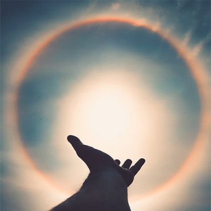 Silhouetted hand reaching toward a glowing sun halo in a cloudy sky
