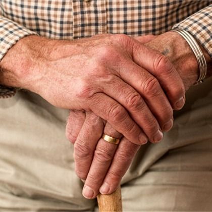 Hands clasped over a wooden cane, with a gold ring and plaid shirt sleeves visible.