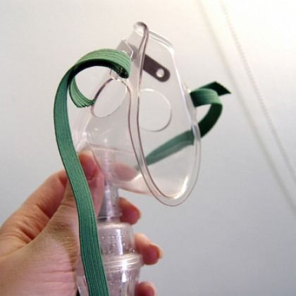 Hand holding a clear oxygen mask with green elastic straps against a white background