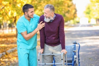 Man in teal scrubs helping an older man with a walker outdoors on a sunny path