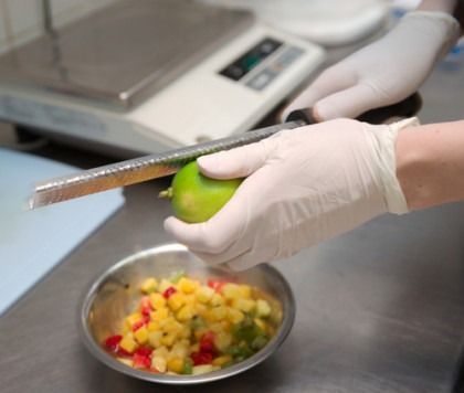 Gloved hands slicing a green mango over a bowl of chopped fruit in a kitchen