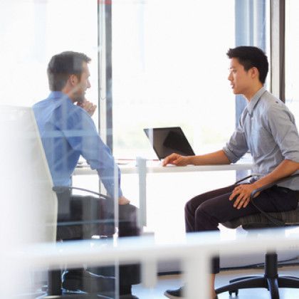 Two men in a bright office meeting, one holding a laptop while they sit across a table.