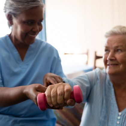Caregiver helps an older adult exercise with a pink dumbbell in a bright room