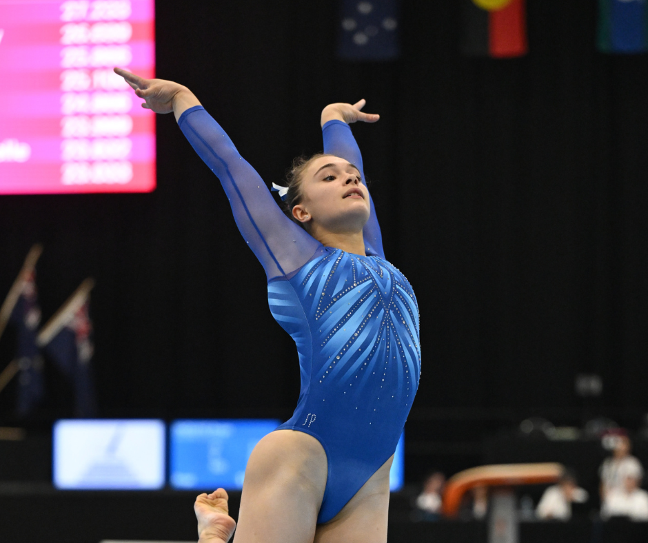 Gymnast in blue leotard, arms raised, mid-routine on floor. Competition setting, focused expression.