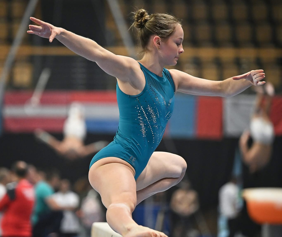 Gymnast in teal leotard balances on beam, arms outstretched, indoor competition setting.