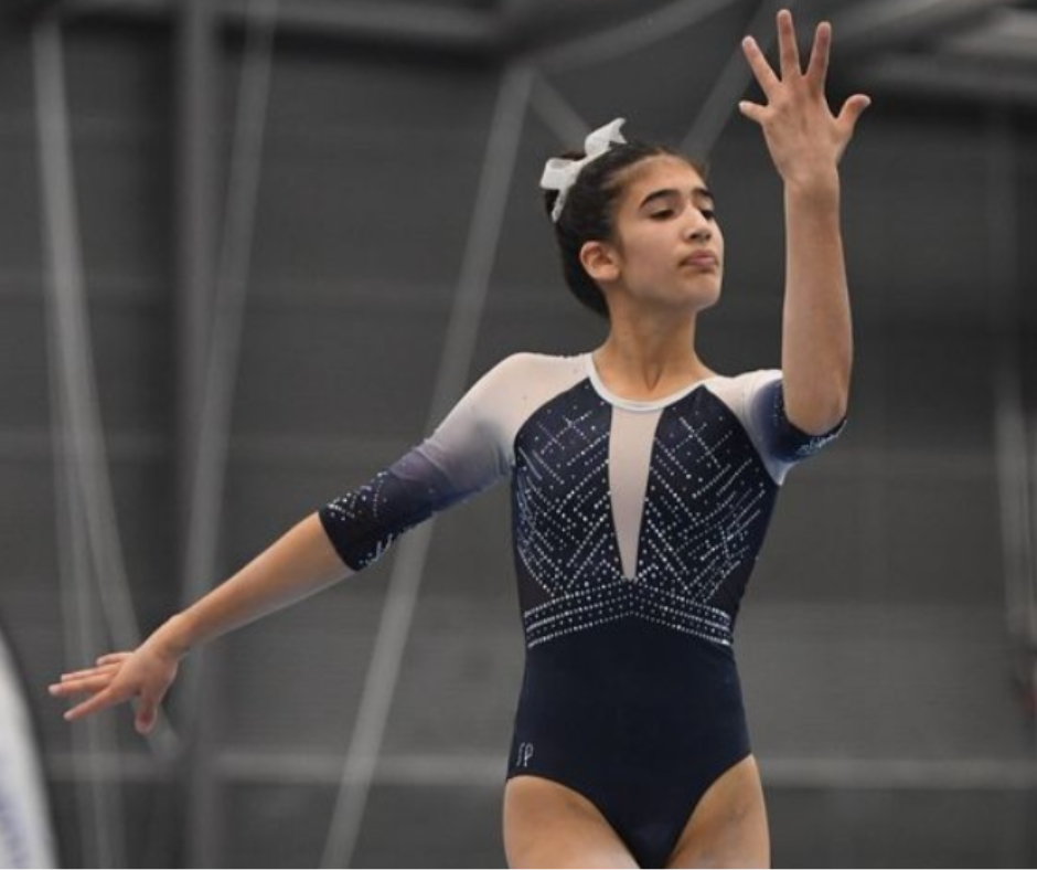 Gymnast in blue leotard performs, arms extended, indoor setting.