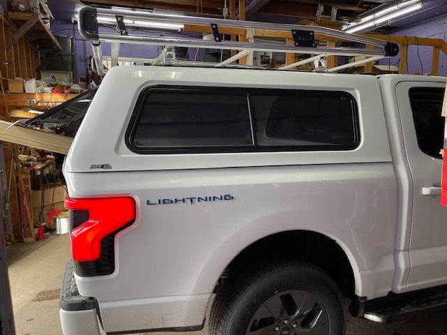 White Ford Lightning truck with a camper shell and ladder rack. Tail light glows red. 