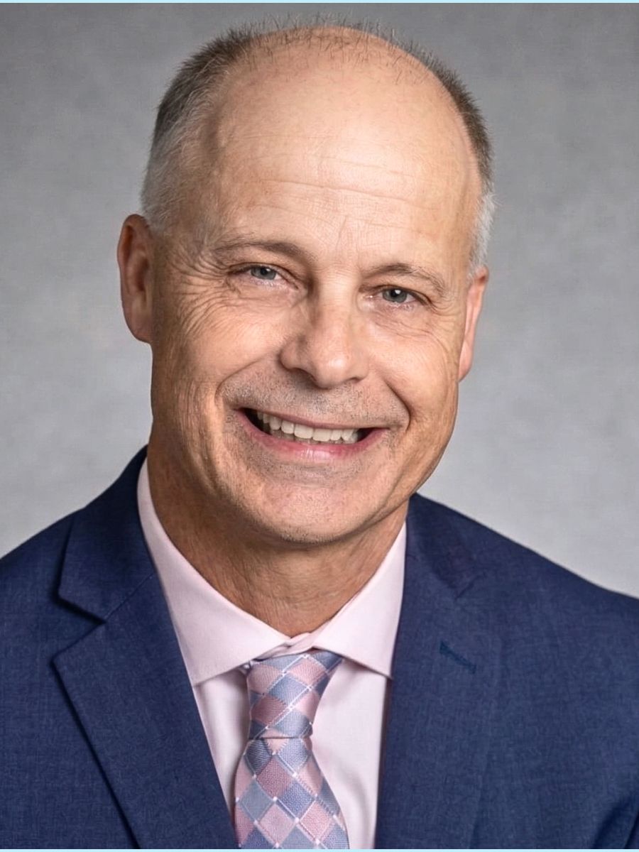 Smiling man in a navy suit and pink tie against a gray studio background