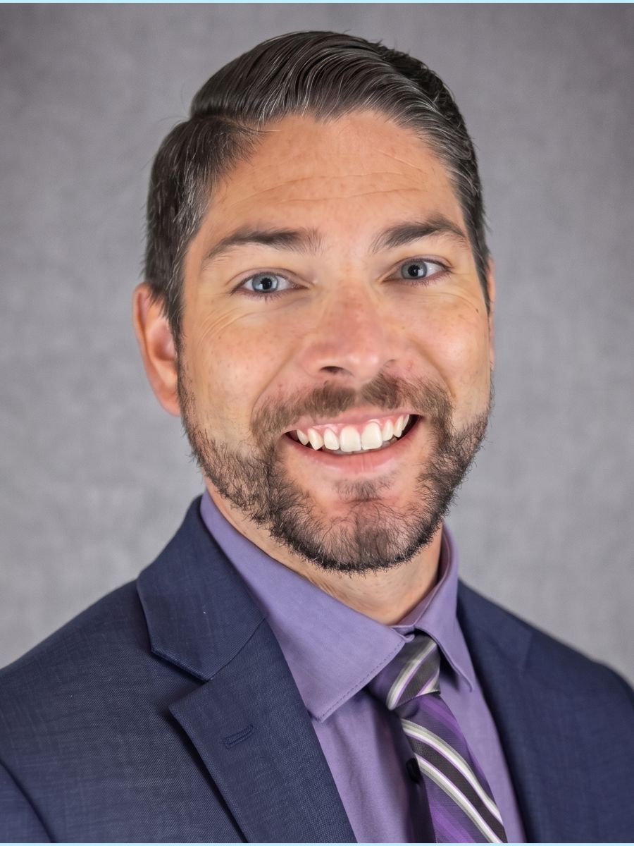 Smiling man in a navy suit, purple shirt, and striped tie against a gray background