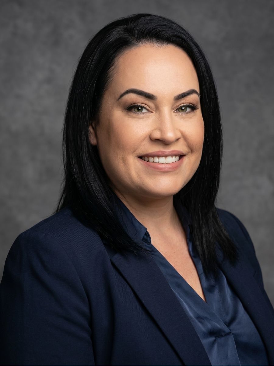 Professional headshot of a smiling person in a navy blouse and blazer against a gray backdrop