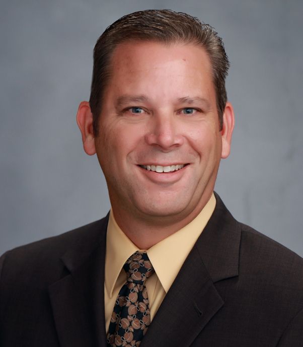 Smiling man in a dark suit, yellow shirt, and patterned tie against a gray backdrop