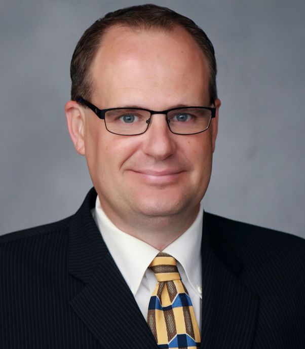 Headshot of a man in glasses, dark suit, and patterned tie against a gray background