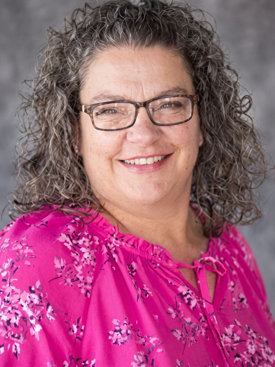 Smiling person with curly gray hair and glasses wearing a bright pink floral blouse against a gray background