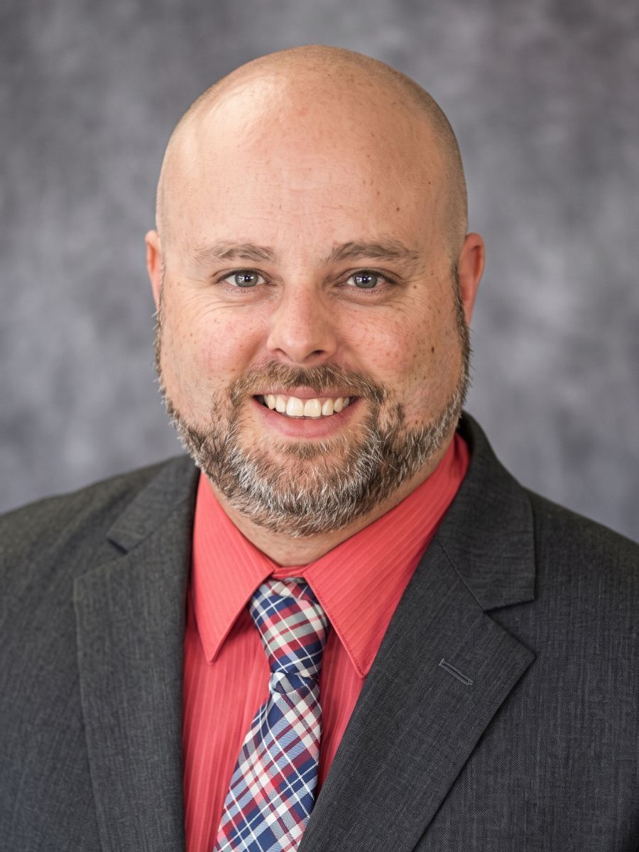 Smiling bald man in a dark suit, red shirt, and plaid tie against a gray background