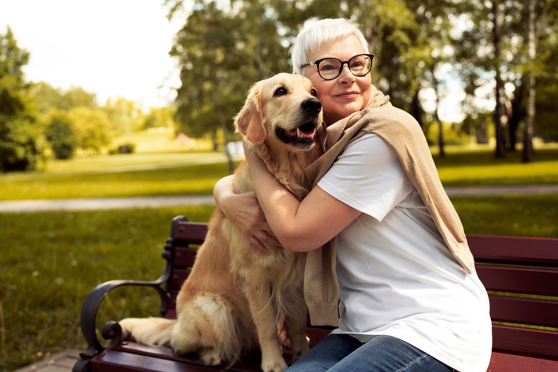 Woman hugging a golden retriever on a park bench in a sunny park