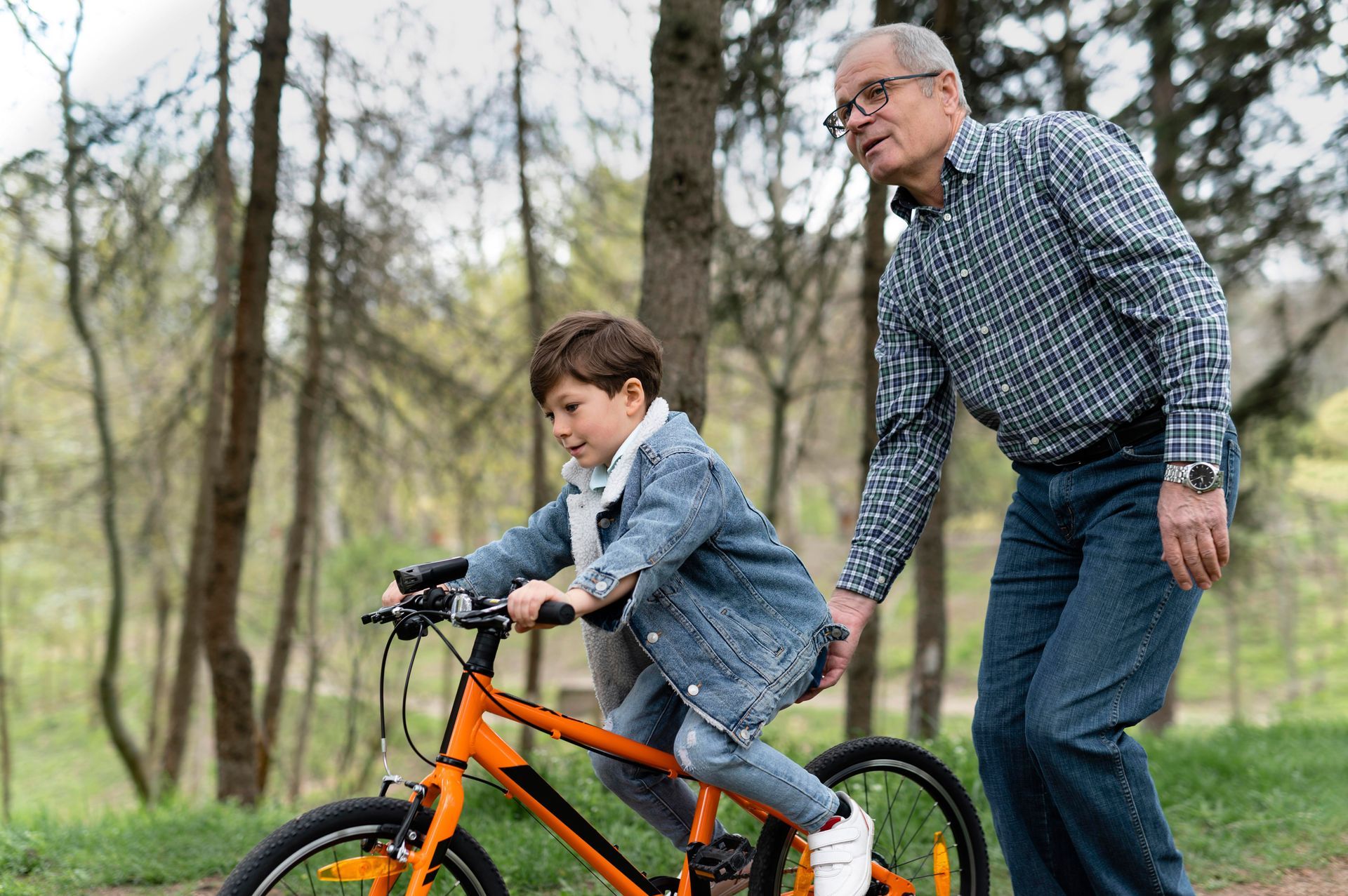 Boy riding orange bicycle while older man walks beside him in a park