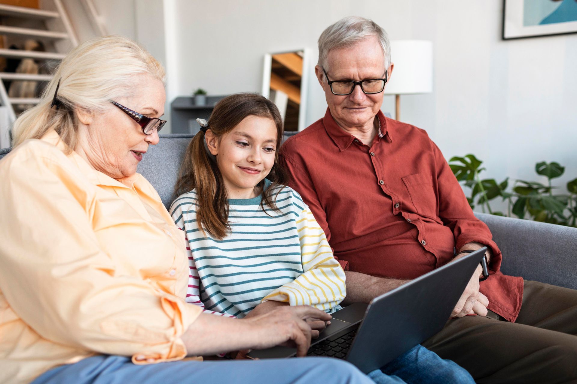 Three people sit on a couch, smiling and looking at a laptop together in a living room.