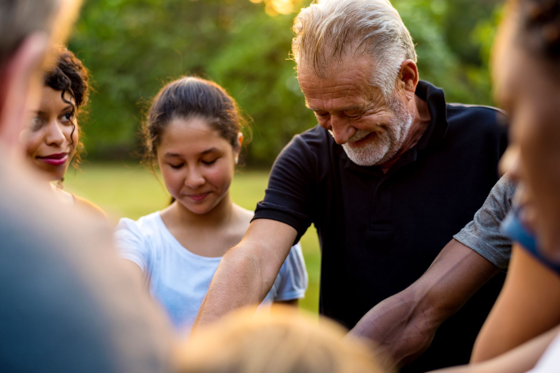 Small group outdoors in a circle, one older man in a black shirt looking down with others around him.