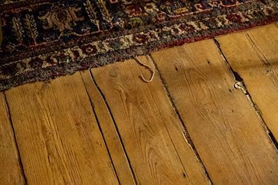 Close-up of aged, wooden floorboards with a patterned rug edge. A necklace rests on the floor.