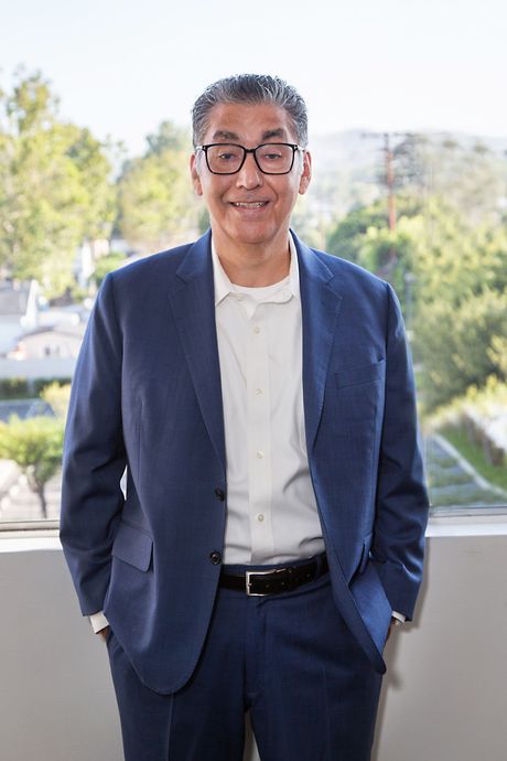 A man in a suit and glasses is standing in front of a window.