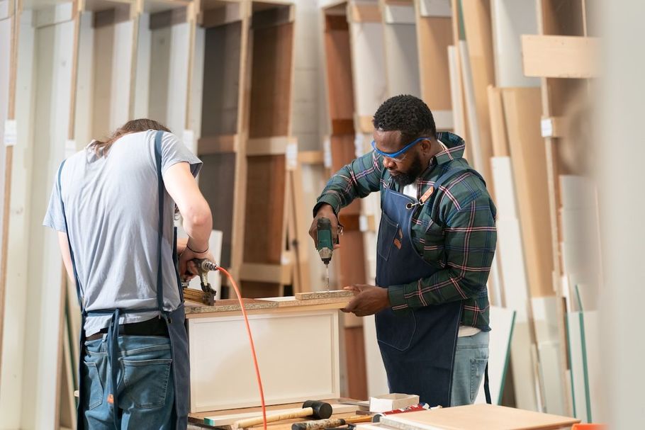 Two workers measuring a wooden board in a workshop, with tools and lumber in the background.