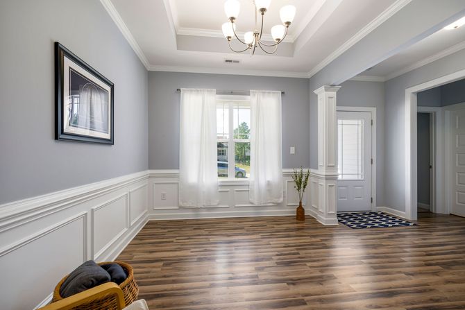 Bright empty hallway with gray walls, white trim, wood floors, chandelier, and a window with a vase of flowers.