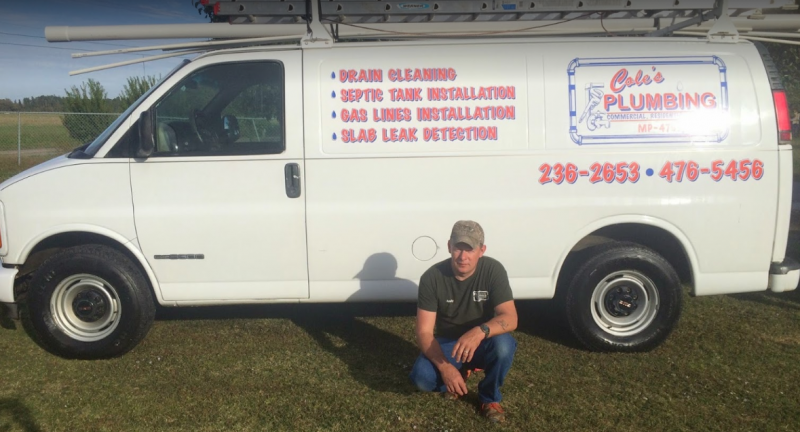 White plumbing service van with red lettering parked on grass; man crouches beside it.