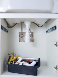 Under-sink cabinet with exposed plumbing and a black toolbox full of cleaning supplies