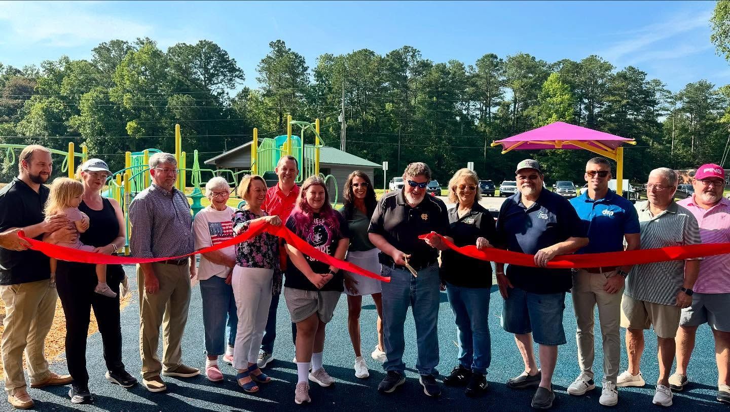 People cutting a red ribbon at a playground dedication ceremony, with a colorful playground in the background.