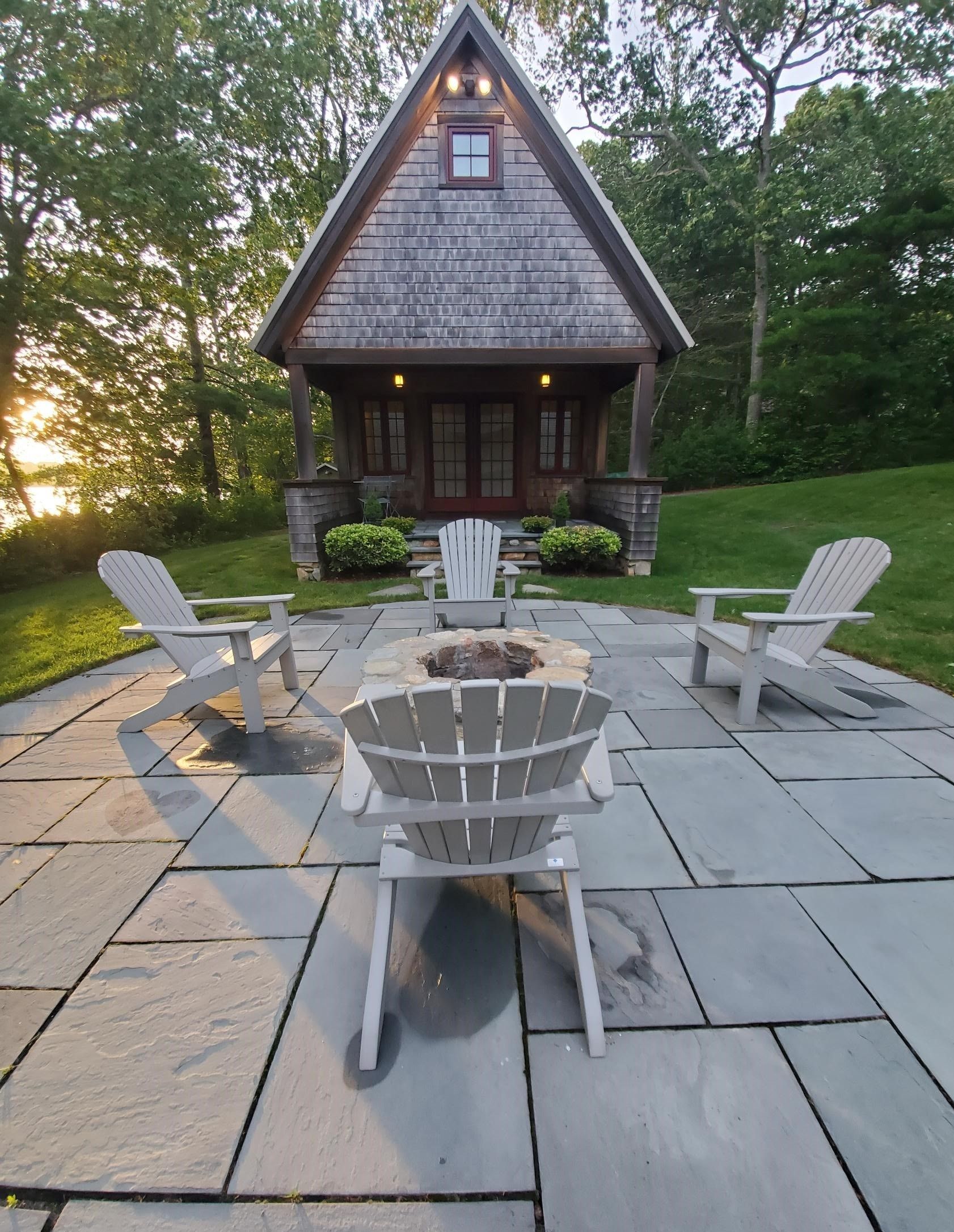Stone patio with Adirondack chairs facing a fire pit and small wooden cottage.