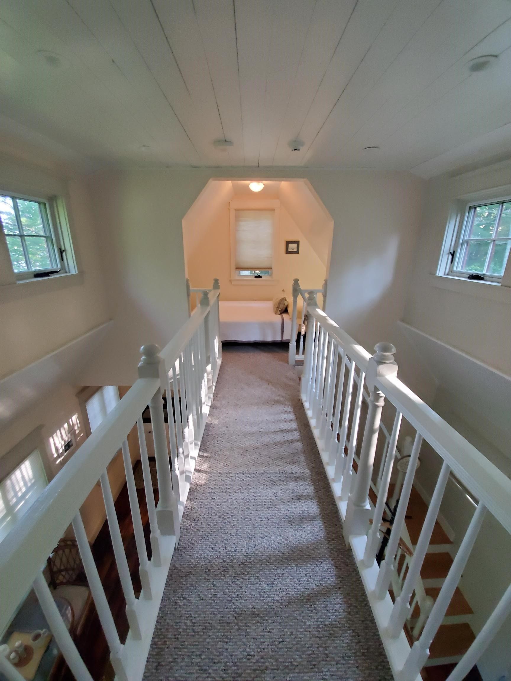 Interior balcony hallway with white railings, carpet, and windows. A bed is visible at the end.
