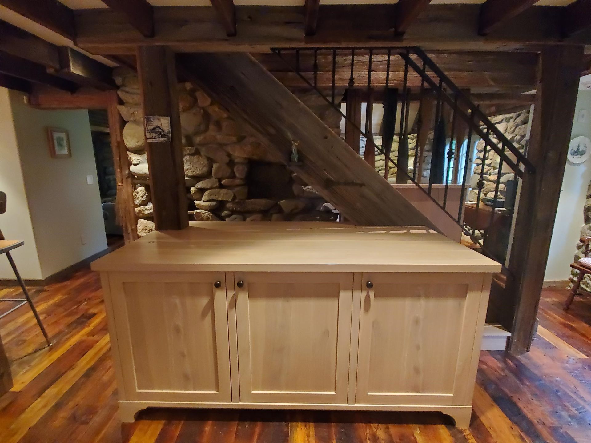 Wooden cabinet with three doors in front of a staircase, wooden beams overhead and stone wall.