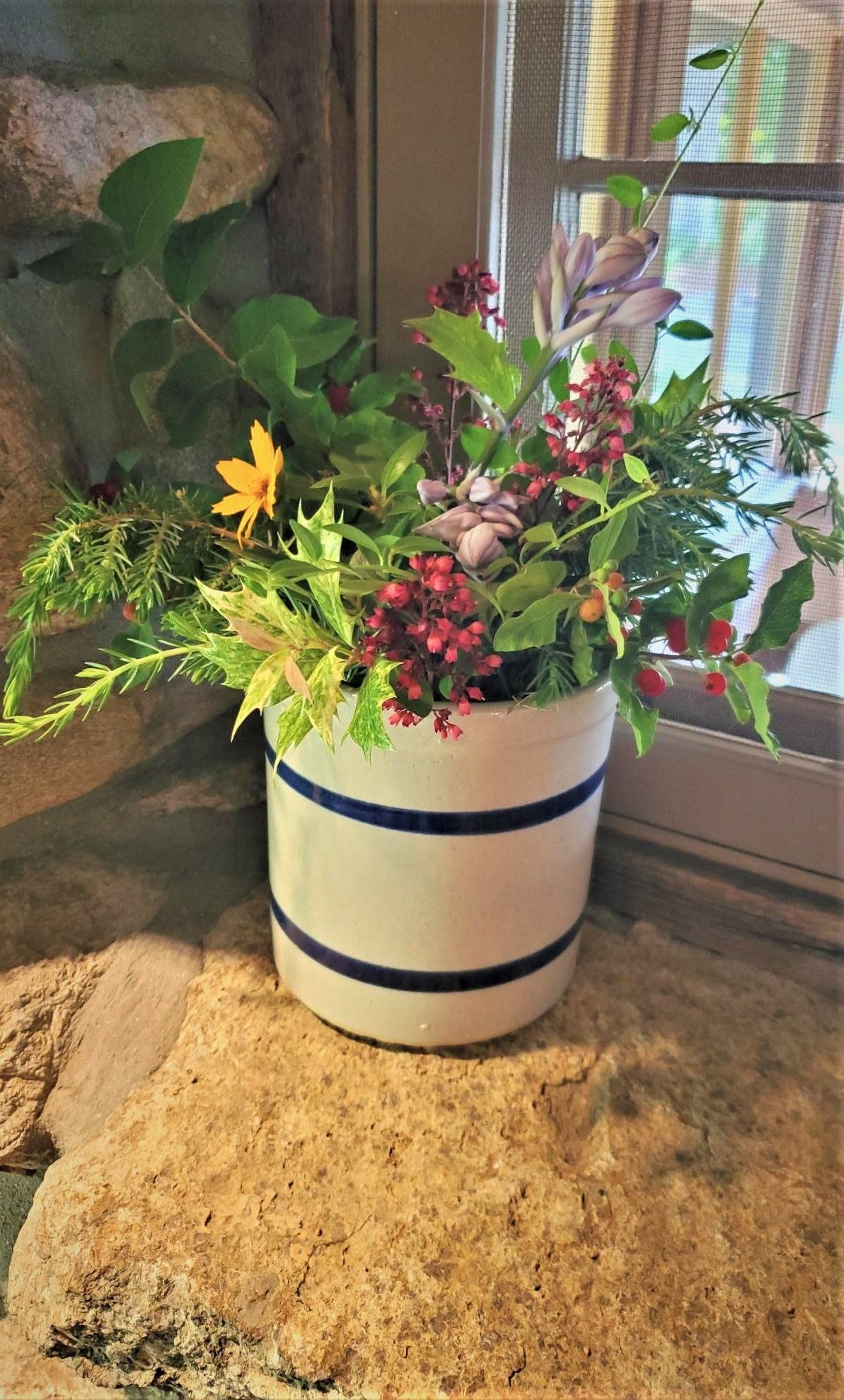 A white ceramic crock with blue stripes filled with mixed foliage and berries on a stone ledge.
