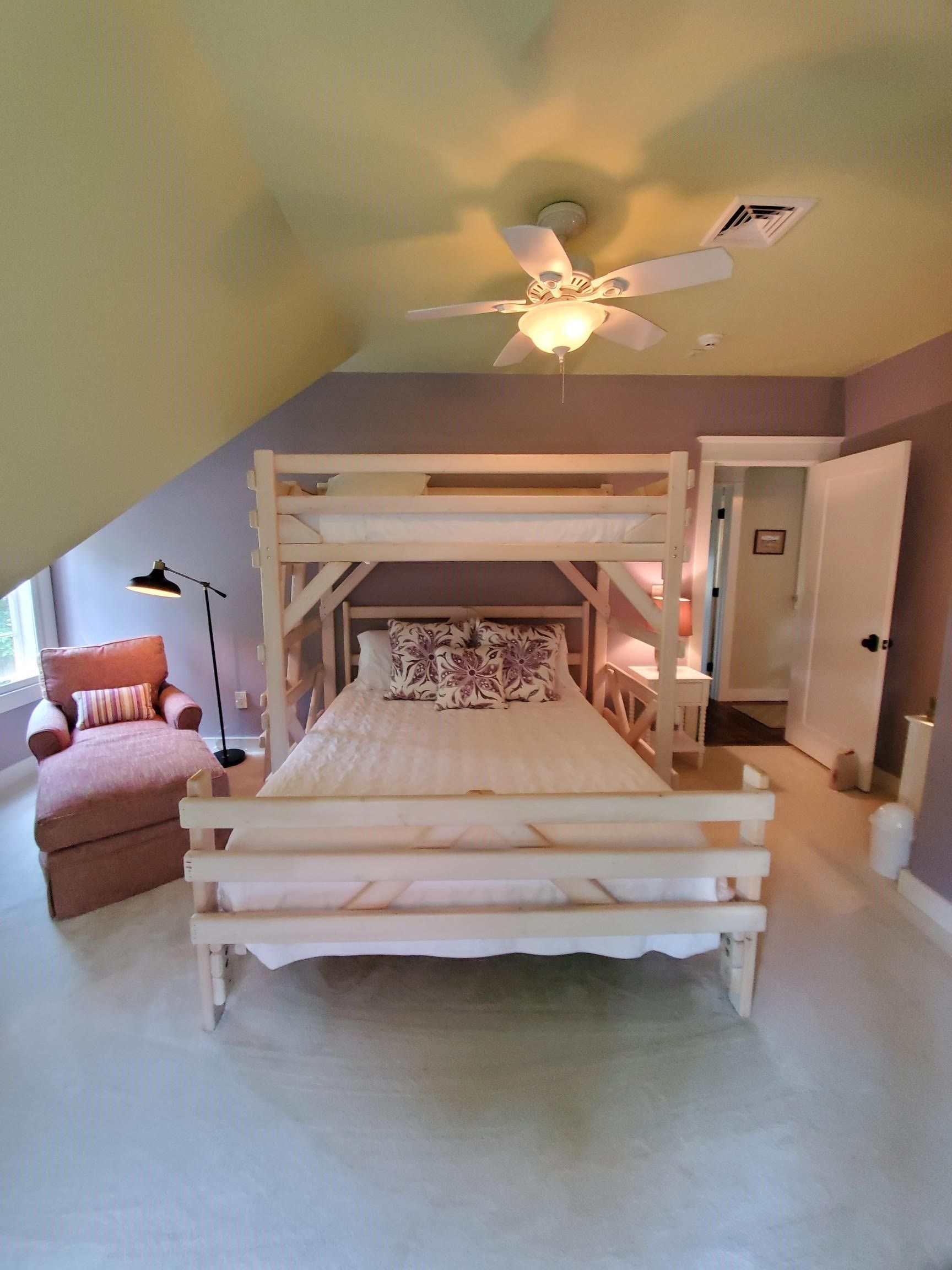 Bedroom with light wood bunk bed, chaise lounge, and door to bathroom. Purple and pale yellow walls, white ceiling fan.