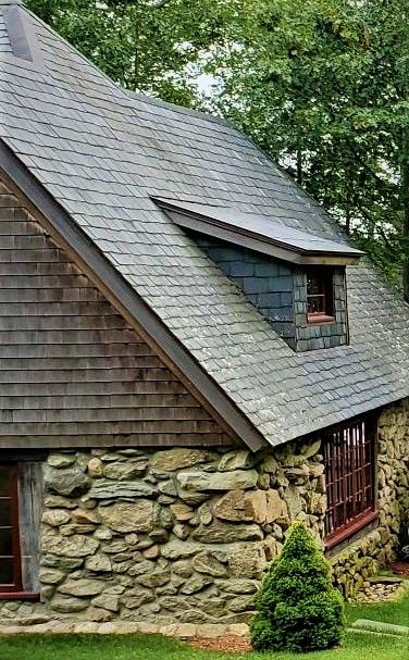 Stone and wood cottage with gray shingle roof and small dormer, set amidst greenery.