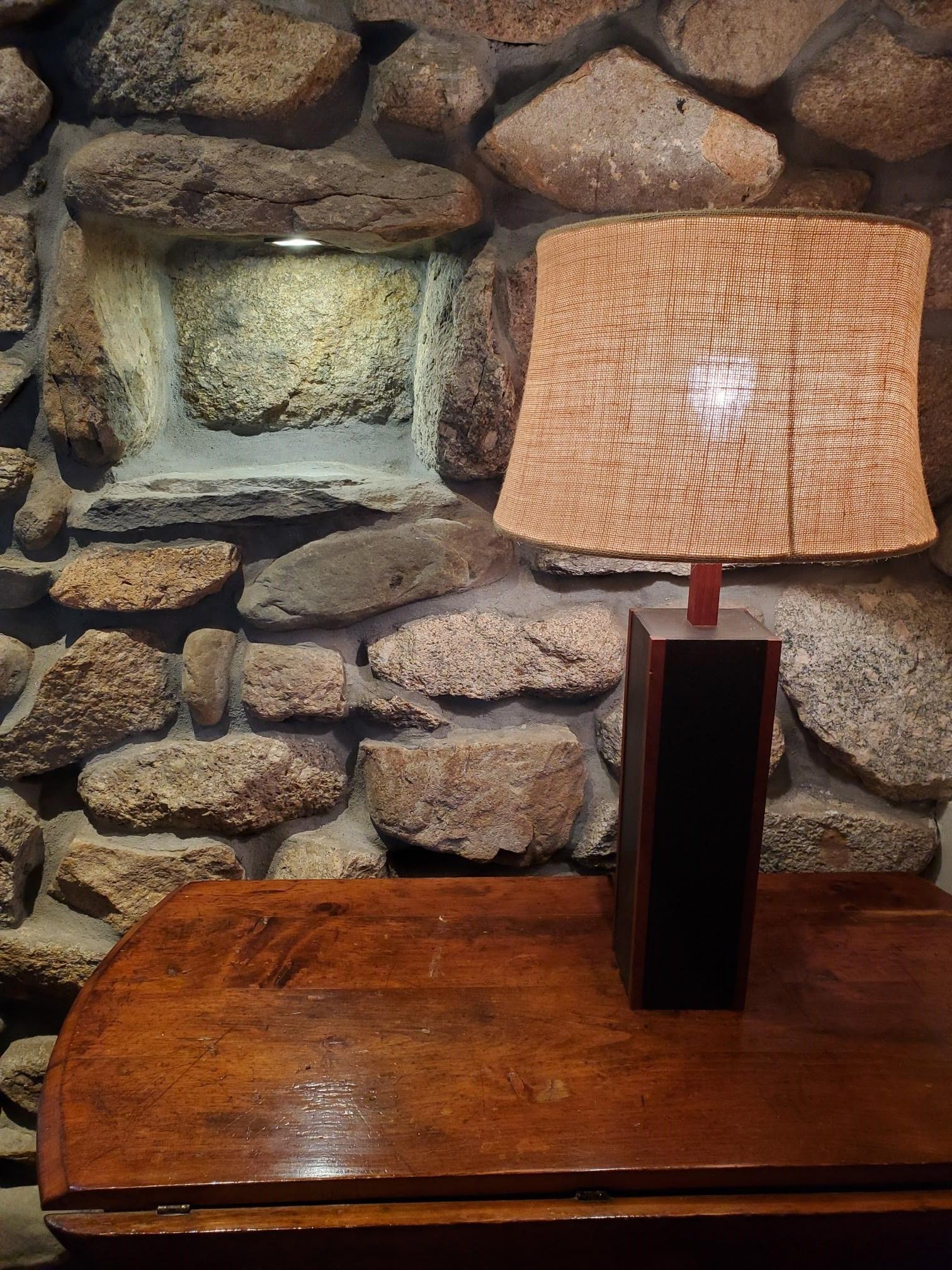 Lamp with a textured beige shade on a wooden table, in front of a stone wall.
