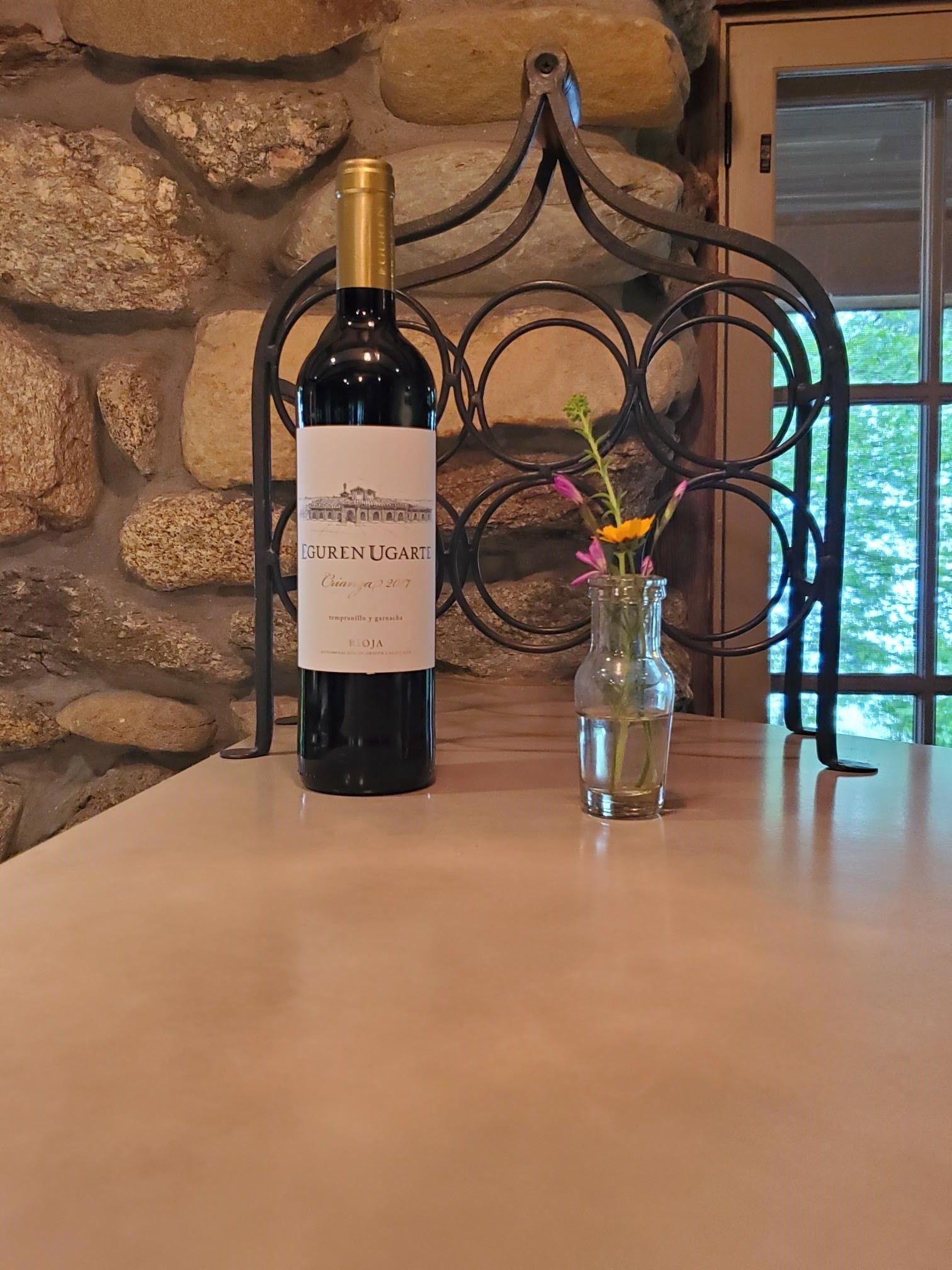 A bottle of red wine and a small vase of flowers on a table, with a stone wall background.