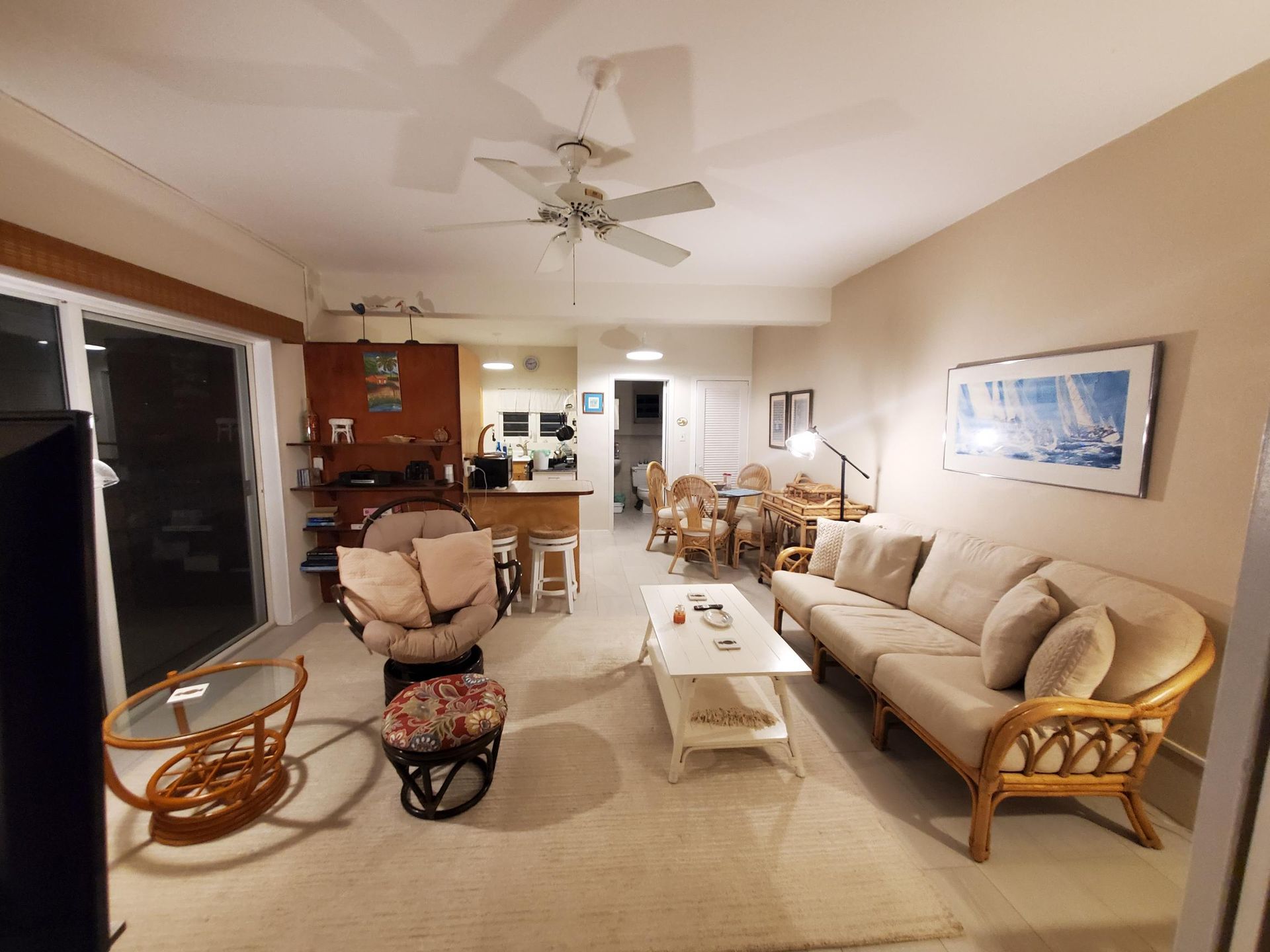Living room with a sofa, chairs, table, and kitchen in the background. Beige walls, warm lighting.