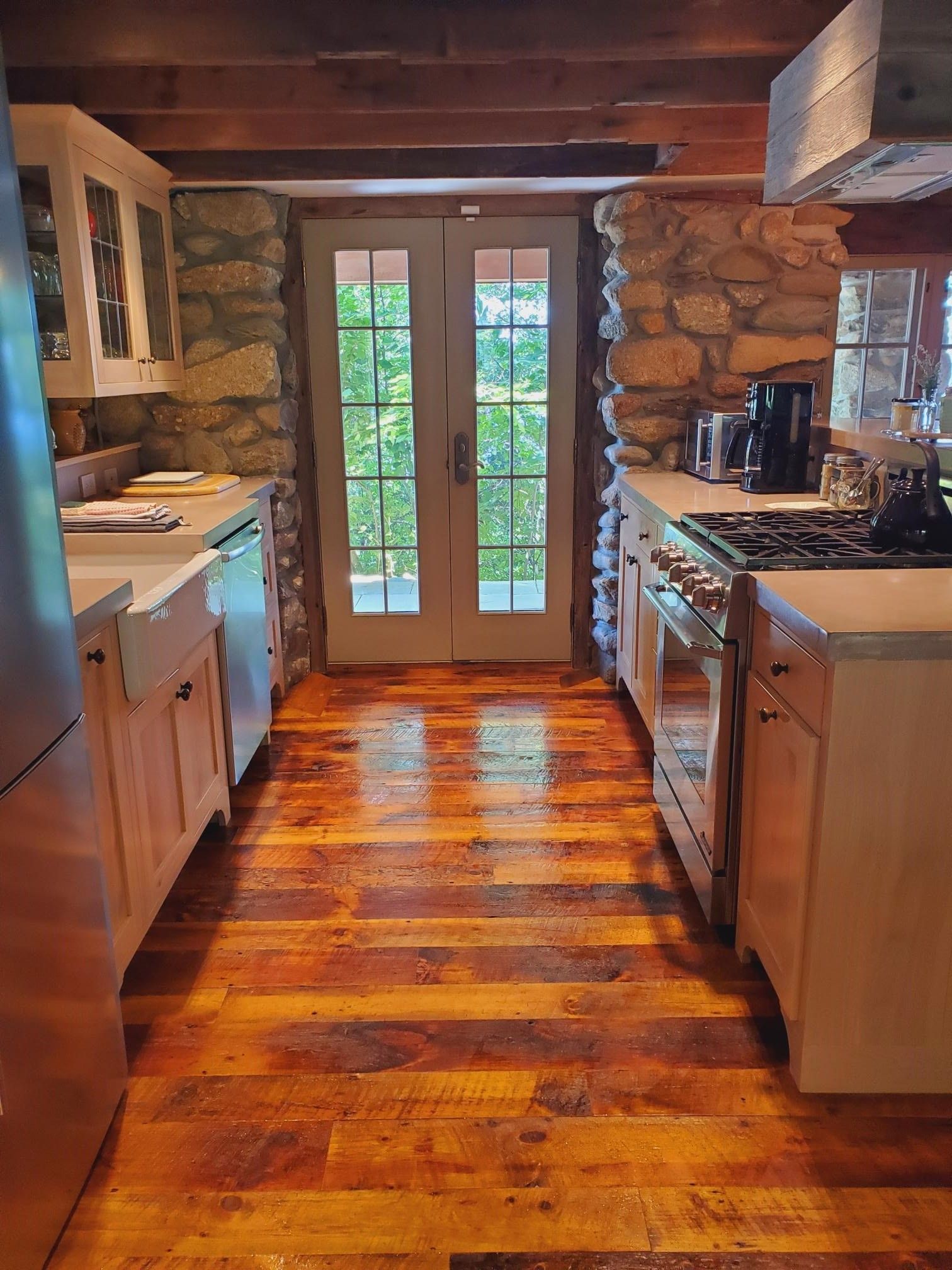 Rustic kitchen with wood floor and stone walls, French doors open to greenery.