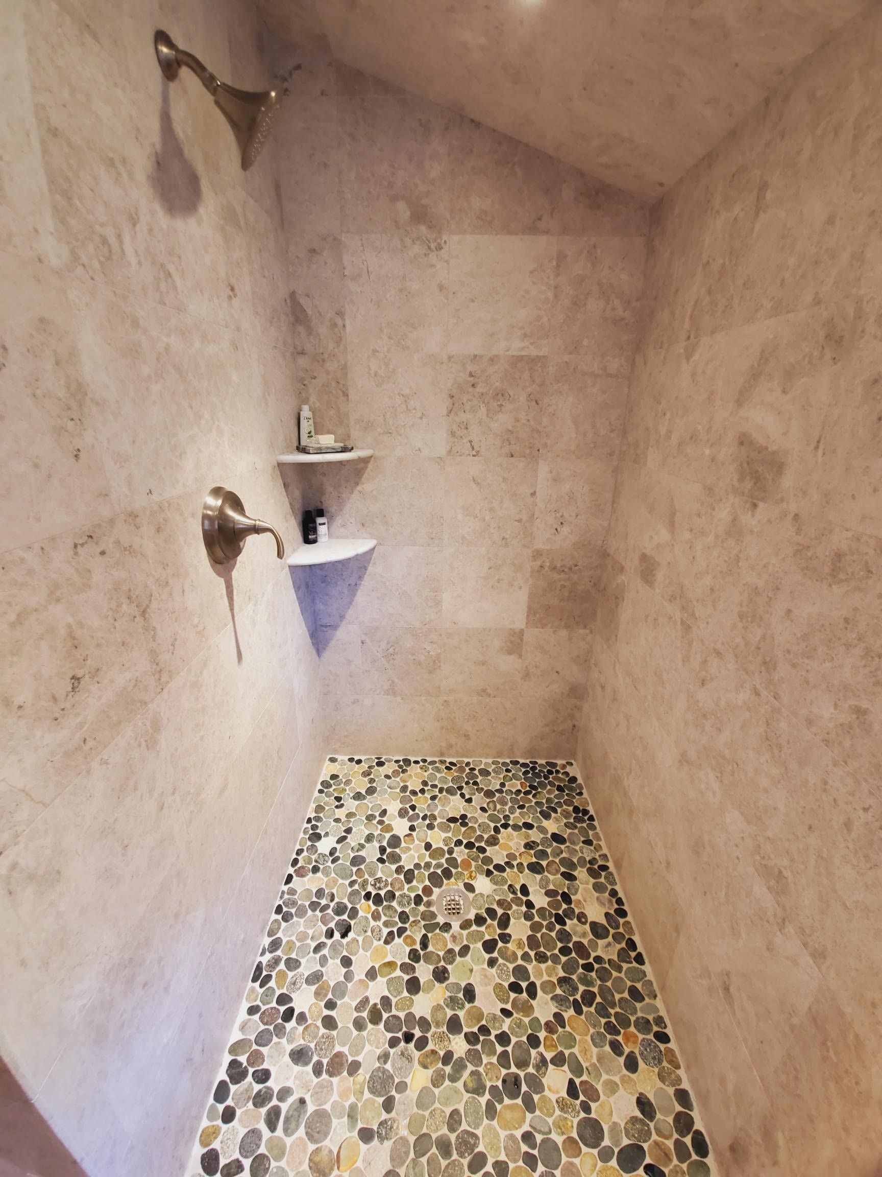 Shower with pebble floor and stone tile walls. Two white triangular shelves hold toiletries.
